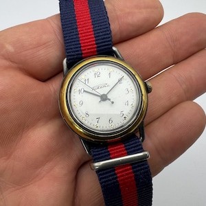 Peut inclure: Une montre-bracelet vintage avec un cadran blanc, une lunette dorée et un bracelet en nylon rayé bleu marine et rouge. Le cadran affiche l'heure avec des chiffres et des aiguilles noirs. Un accessoire classique.