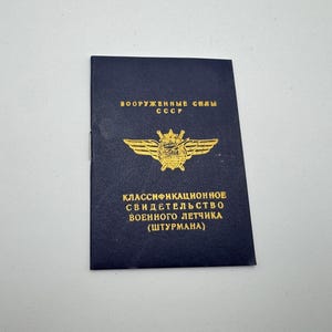 May include: A navy blue document with gold lettering and a gold emblem featuring wings and a star. The text reads "Armed Forces USSR" and "Classification Certificate of Military Pilot (Navigator)."