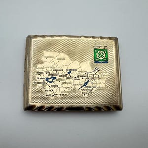 Könnte beinhalten: Eine rechteckige, goldfarbene Zigarettenetui mit detailliertem Kartendesign. Die Karte zeigt verschiedene Städte und geografische Details, mit einem grünen und blauen Emblem oben rechts. Die Hülle hat einen strukturierten Rand.