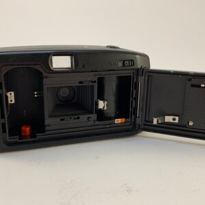 May include: A black and silver 35mm film camera with the back open, revealing the film compartment and lens. The camera has a built-in flash and a small LCD screen.