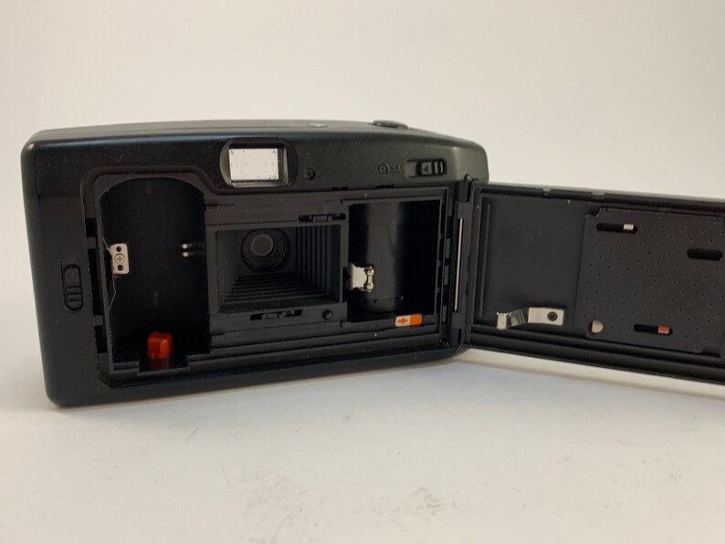 May include: A black and silver 35mm film camera with the back open, revealing the film compartment and lens. The camera has a built-in flash and a small LCD screen.