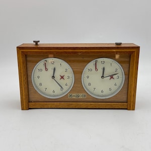 May include: A rectangular wooden analog chess clock with two circular white clock faces featuring black numerals from 1 to 12. Each face has black hour and minute hands, a red second hand, and a red star icon. The brand name GARDE is printed in black.