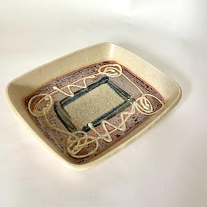 May include: A rectangular ceramic tray with a beige rim and a textured interior. The interior features a central rectangle in beige and blue, surrounded by brown and red accents, and white squiggly lines.