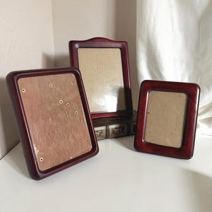 May include: Three dark cherry-toned wooden picture frames of varying sizes. The left frame shows a light brown backing with small metal pins, the center frame is elevated on a book, and the right frame is smaller with a plain tan insert.