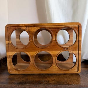 May include: A rectangular wooden wine rack featuring a natural brown grain finish. The structure includes six circular cutouts arranged in two rows of three, designed to hold bottles horizontally. The frame has a smooth, polished surface.