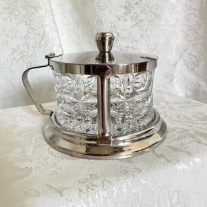 May include: A clear glass condiment jar with a geometric pressed pattern, encased in a polished silver-tone metal frame. The jar features a matching silver-tone metal lid with a central knob handle and a side handle attached to the frame.