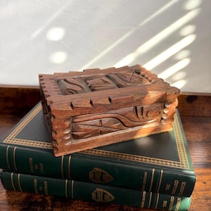 May include: A rectangular wooden box with a dark brown finish and intricate hand-carved geometric patterns on the lid and sides. The box features a notched, serrated edge design along the top perimeter and rests on two dark green hardcover books.