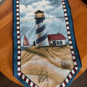 May include: A rectangular table runner featuring a woven scene of a blue and white striped lighthouse, a small house with a red roof, a sailboat on the water, and sandy dunes. The runner has a blue border with a red and white checkered pattern.