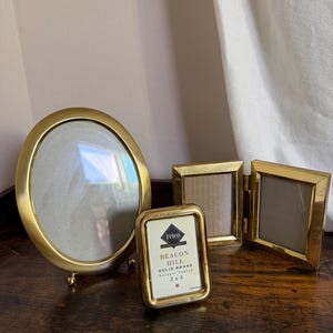 May include: A collection of three gold-toned metal picture frames. One is a large oval frame with small feet, one is a small rectangular frame with a label reading Fetco International Beacon Hill Solid Brass 2 x 3, and one is a double hinged frame.