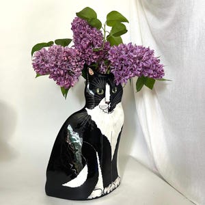 May include: A black and white cat-shaped ceramic vase holding a bouquet of purple lilacs. The vase has green eyes and a white chest. The flowers are a vibrant purple with green leaves, creating a striking contrast.