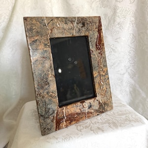 May include: A rectangular picture frame featuring a faux marble finish with a blend of dark brown, tan, and grey tones. The surface displays intricate veining patterns in white and deep red, creating a textured appearance around the central glass.