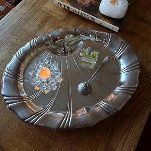 May include: A large, oval, silver-toned metal tray with a scalloped edge and radial grooves. On the tray sits a clear glass candle holder with a lit tea light, a small square glass perfume bottle, and a long-handled silver-toned metal spoon.