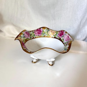 May include: A white porcelain bowl with three small feet and a scalloped rim. The interior edge features a decorative band of pink and yellow roses set against a gold-painted border. The bowl has a smooth, glossy finish and a deep, rounded basin.