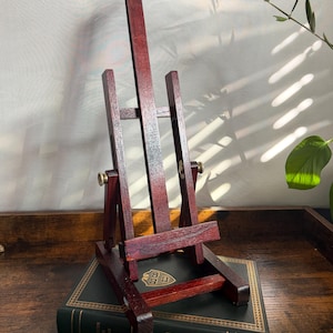 May include: A dark reddish-brown wooden tabletop easel featuring a central vertical support, two side supports, and an adjustable horizontal ledge. The easel includes two metallic knobs on the sides for adjusting the angle and height of the frame.