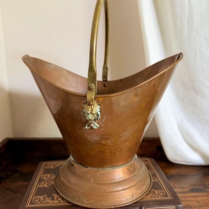 May include: A copper coal scuttle with a wide, flared rim and a circular base. The vessel features a tall, arched brass handle attached to the sides with decorative brass lion head mounts. The metal surface shows a warm, aged patina throughout.