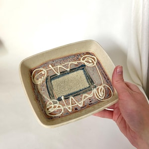 May include: A square ceramic dish with a beige exterior and a rectangular design in the center. The design features a beige rectangle with blue and brown accents, and white swirls. The dish is held by a hand.