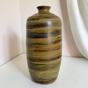 May include: A cylindrical ceramic vase featuring a mottled, earthy color palette of olive green, dark brown, and tan. The surface displays horizontal, layered bands of color with a smooth, matte finish, a narrow neck, and a small, circular opening.