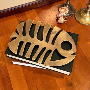 May include: A brass fish skeleton-shaped object rests on a stack of books with a dark green cover. The fish skeleton is a warm gold color and has a circular eye. The books are on a wooden surface.