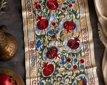 Puede incluir: Un camino de mesa rectangular de tela color hueso con bordados intrincados de granadas rojas, enredaderas verdes y detalles florales azules. El borde presenta un patrón de espiral continuo en hilo rojo, azul y amarillo.