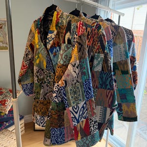 May include: A collection of three quilted jackets hanging on a metal rack. Each jacket features a patchwork design composed of various fabric squares in vibrant colours including yellow, blue, red, and green, with intricate patterns and stitching.