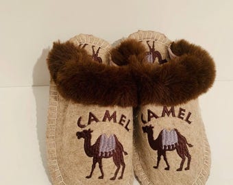 Può includere: Un paio di pantofole in feltro beige con finiture in finta pelliccia marrone. Ogni pantofola presenta un disegno di cammello ricamato con la scritta "CAMEL" sopra. Le pantofole hanno un bordo cucito.