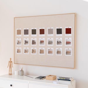 Pode incluir: Um quadro emoldurado com amostras de tecido e pequenas fotos de móveis. As amostras são em vários tons de castanho, cinza e vermelho. O quadro está montado numa parede branca acima de uma cómoda branca. Uma lâmpada de madeira e um manequim de artista em madeira também são visíveis.