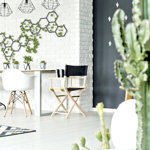 May include: A white dining table with four chairs, including a black director's chair, in a modern living room with a white brick wall and a black wall with cactus decals. A hexagonal metal wall decor with green plants is on the white brick wall.