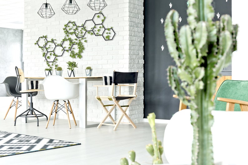 May include: A white dining table with four chairs, including a black director's chair, in a modern living room with a white brick wall and a black wall with cactus decals. A hexagonal metal wall decor with green plants is on the white brick wall.
