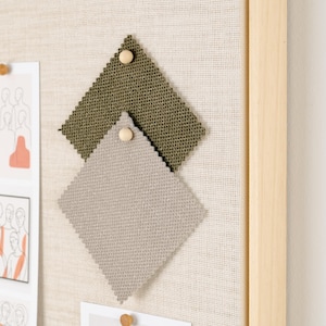 May include: A neutral-toned fabric-covered bulletin board with a light wooden frame. Two fabric swatches, one olive green and one gray, are pinned to the board. Several sketches and drawings are also pinned to the board with wooden push pins.