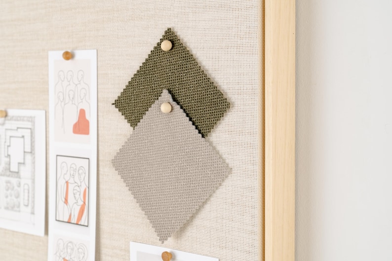 May include: A neutral-toned fabric-covered bulletin board with a light wooden frame. Two fabric swatches, one olive green and one gray, are pinned to the board. Several sketches and drawings are also pinned to the board with wooden push pins.