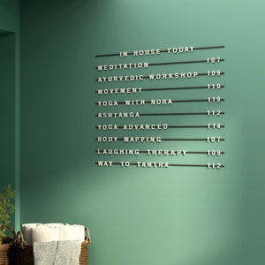 May include: A black and white sign on a green wall lists yoga and wellness class titles, including Meditation, Ayurvedic Workshop, and Yoga with Nora. The sign is mounted on a green wall with a basket and towels in the foreground.
