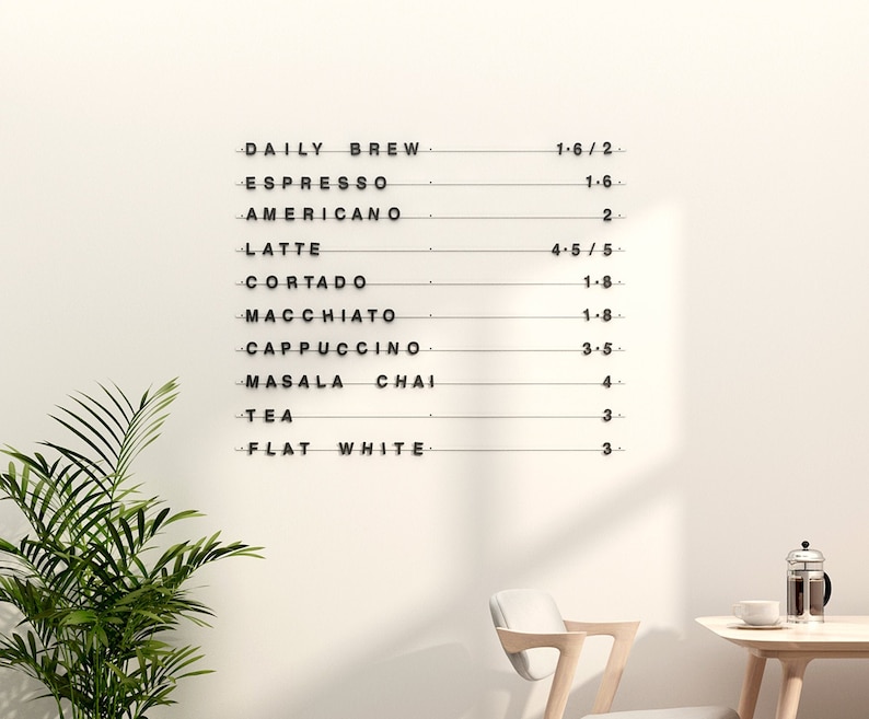 May include: A minimalist coffee menu on a white wall, listing various coffee drinks such as Espresso, Latte, and Cappuccino. The menu is in black lettering with prices listed next to each item. A green plant and a small table with a chair are in the foreground.