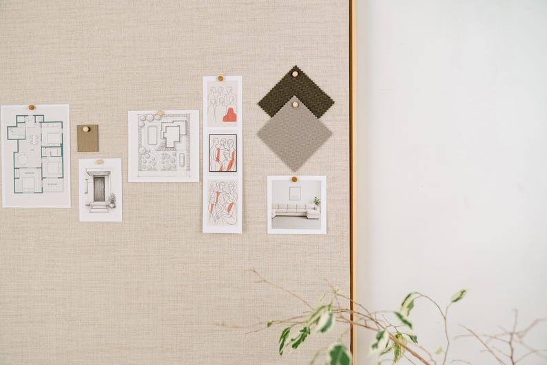 May include: A neutral-toned mood board with architectural sketches, fabric swatches, and art prints. The board is pinned with wooden push pins. A sprig of greenery is in the foreground.