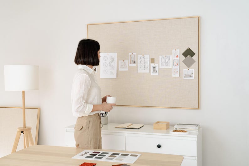 May include: A woman stands near a large, beige fabric-covered bulletin board with various architectural plans and color swatches pinned to it. A white lamp and a wooden table are in the foreground. The scene suggests an interior design or architectural planning space.