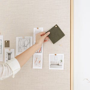 May include: A woman is holding a square, olive-green fabric swatch pinned to a beige bulletin board. The board also displays architectural sketches and a photo of a living room. The scene suggests interior design or home decor planning.