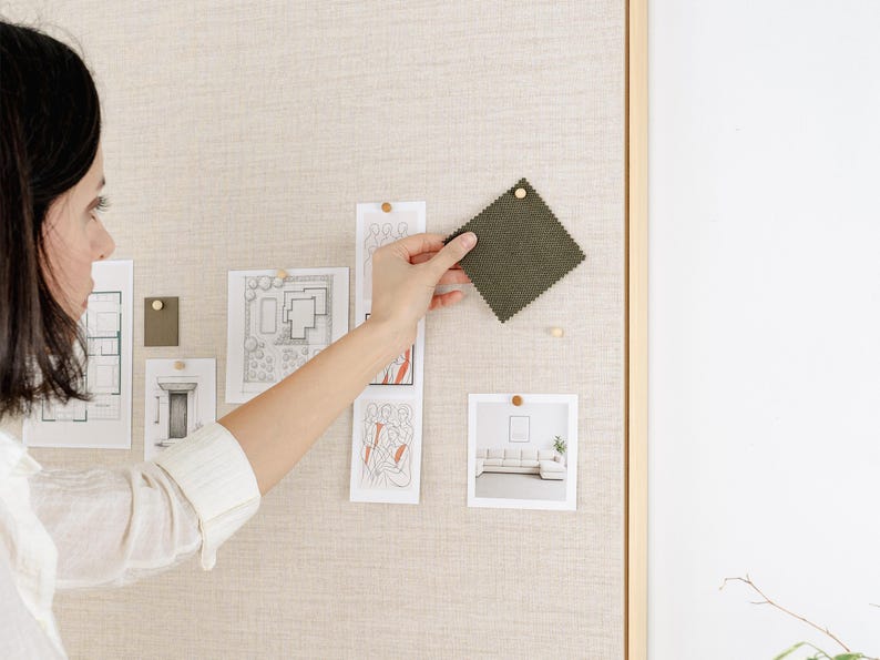 May include: A woman is holding a square, olive-green fabric swatch pinned to a beige bulletin board. The board also displays architectural sketches and a photo of a living room. The scene suggests interior design or home decor planning.