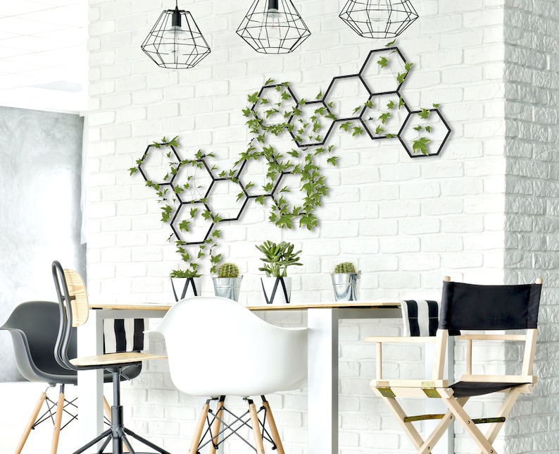 May include: A black metal hexagonal wall decor with green ivy plants growing through it. The decor is hanging on a white brick wall. There are three black pendant lights hanging from the ceiling above the decor. There are three chairs and a table in the foreground.