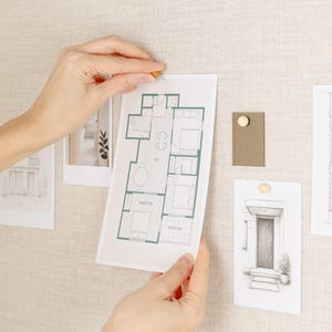 May include: A person's hands are shown pinning a blueprint of a house plan to a beige fabric board. Other architectural sketches and a color swatch are also pinned to the board with wooden push pins. The blueprint is outlined in green.