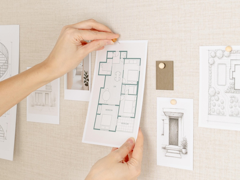 May include: A person's hands are shown pinning a blueprint of a house plan to a beige fabric board. Other architectural sketches and a color swatch are also pinned to the board with wooden push pins. The blueprint is outlined in green.