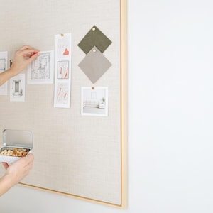 May include: A neutral-toned bulletin board with a wooden frame displays architectural sketches, fabric swatches, and photographs. A small tin filled with nuts is held in front of the board. The board is mounted on a white wall.
