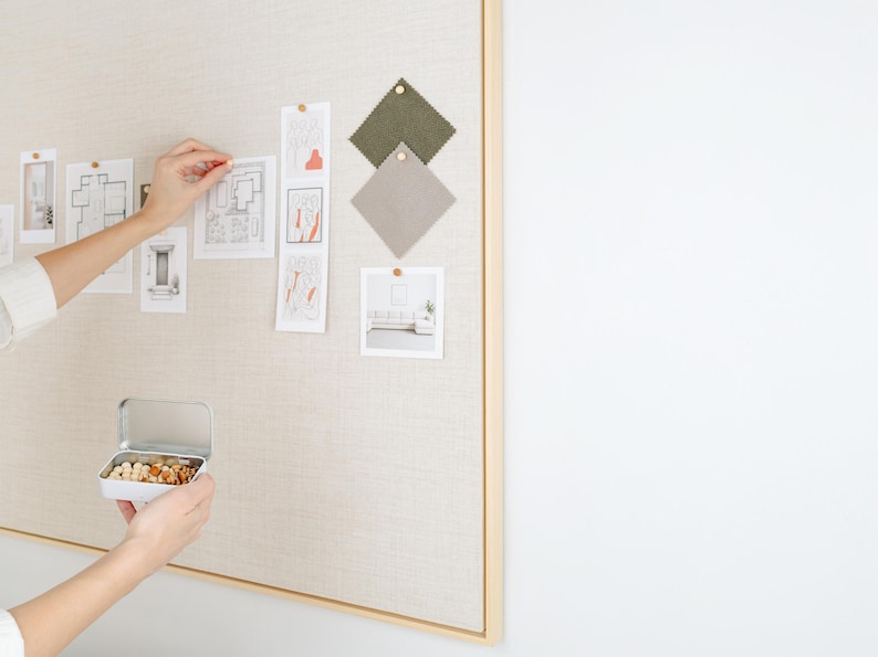 May include: A neutral-toned bulletin board with a wooden frame displays architectural sketches, fabric swatches, and photographs. A small tin filled with nuts is held in front of the board. The board is mounted on a white wall.