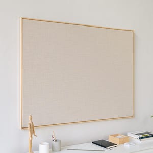 May include: A large, rectangular bulletin board with a light beige fabric surface and a thin, gold-colored frame. The board is mounted on a white wall. A desk with a wooden artist's mannequin, pencils, and a notebook is in front of the board.