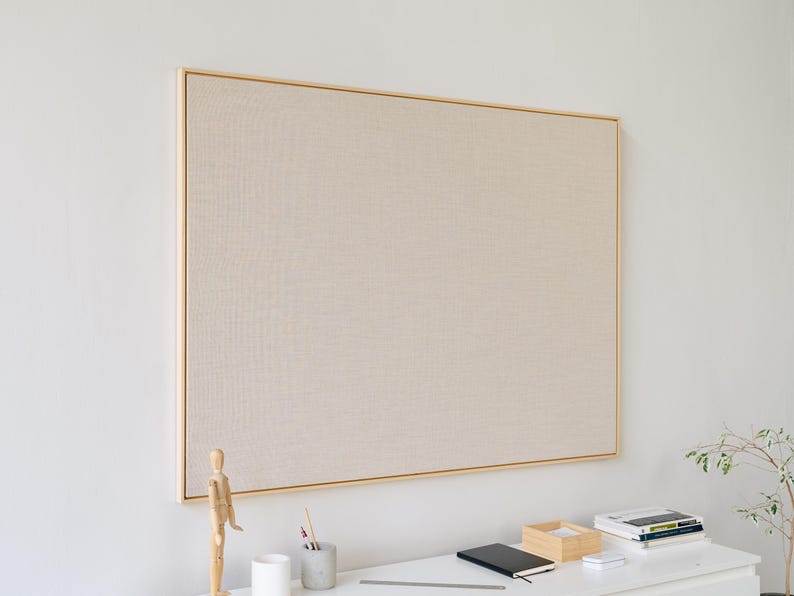May include: A large, rectangular bulletin board with a light beige fabric surface and a thin, gold-colored frame. The board is mounted on a white wall. A desk with a wooden artist's mannequin, pencils, and a notebook is in front of the board.