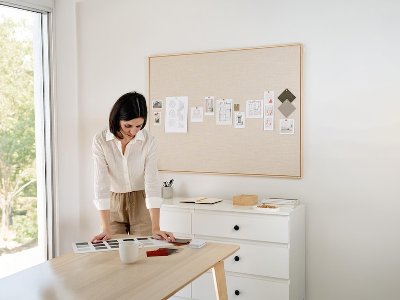 May include: A woman in a white shirt and tan shorts examines color swatches on a light wood table. A large bulletin board with design samples hangs on the wall. A white dresser and a window are also visible.