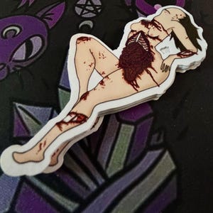 May include: A stack of stickers featuring a stylized illustration of a person with exposed internal organs. The artwork is in shades of tan, red, and black, with a white border. The stickers are stacked on a black surface with purple geometric and occult-themed designs.