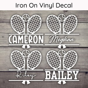 May include: White vinyl decal featuring tennis rackets and balls, with names Cameron, Meghan, Riley, and Bailey. The text "Iron On Vinyl Decal" is at the top. The design is on a wooden background.