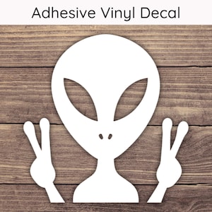 May include: White adhesive vinyl decal featuring an alien with large eyes and peace signs. The text "Adhesive Vinyl Decal" is above the alien. The decal is on a wooden background.