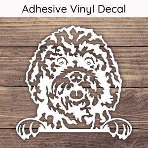 May include: White adhesive vinyl decal featuring a detailed illustration of a dog's face and paws. The text "Adhesive Vinyl Decal" is at the top. The decal is on a wooden background.