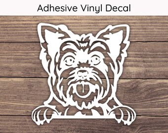 May include: White adhesive vinyl decal of a Yorkshire Terrier dog. The dog's face is detailed with eyes, nose, and tongue. The decal includes the text "Adhesive Vinyl Decal" at the top. The dog's paws are visible at the bottom.