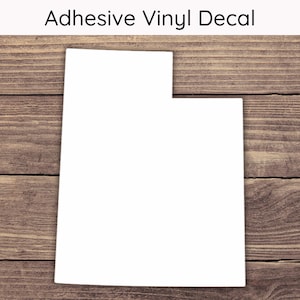 May include: White vinyl decal of the state of Utah. The decal is on a brown wooden background.
